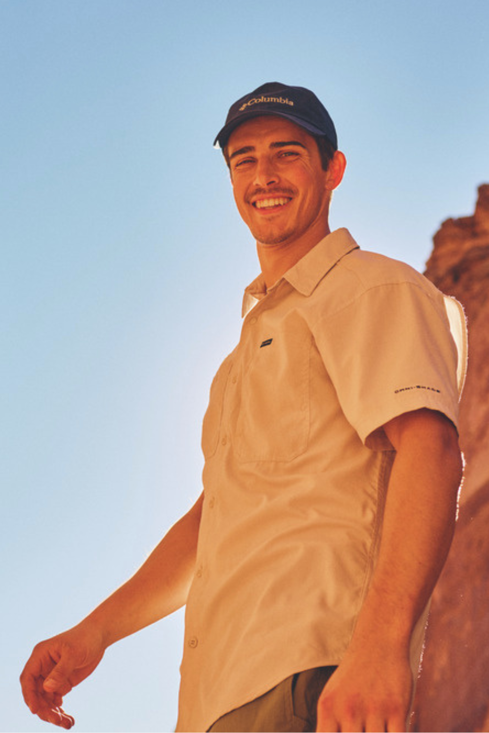 Homme souriant portant une chemise anti-UV beige Columbia, certifiée UPF50+, dans un environnement ensoleillé.